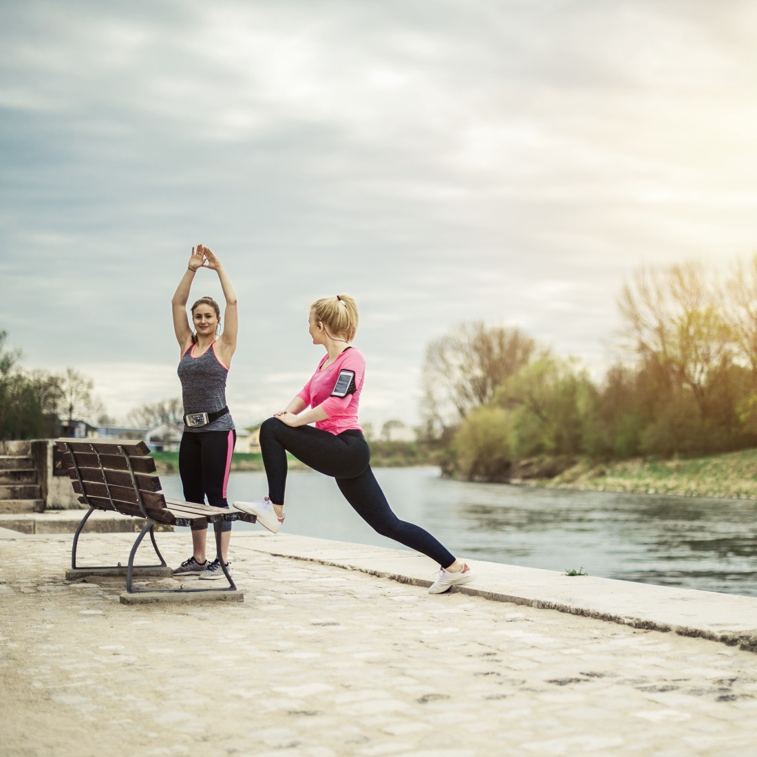 Zwei Frauen trainieren im Freien. Eine dehnt sich, die andere hebt die Arme. Fluss und Bäume im Hintergrund.