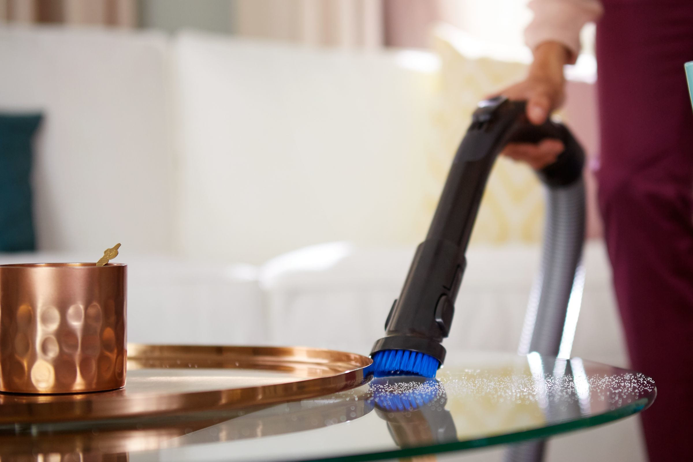 Eine Person saugt einen Glastisch. Eine Kupferbecher und ein Tablett sind sichtbar. Weißes Sofa im Hintergrund.