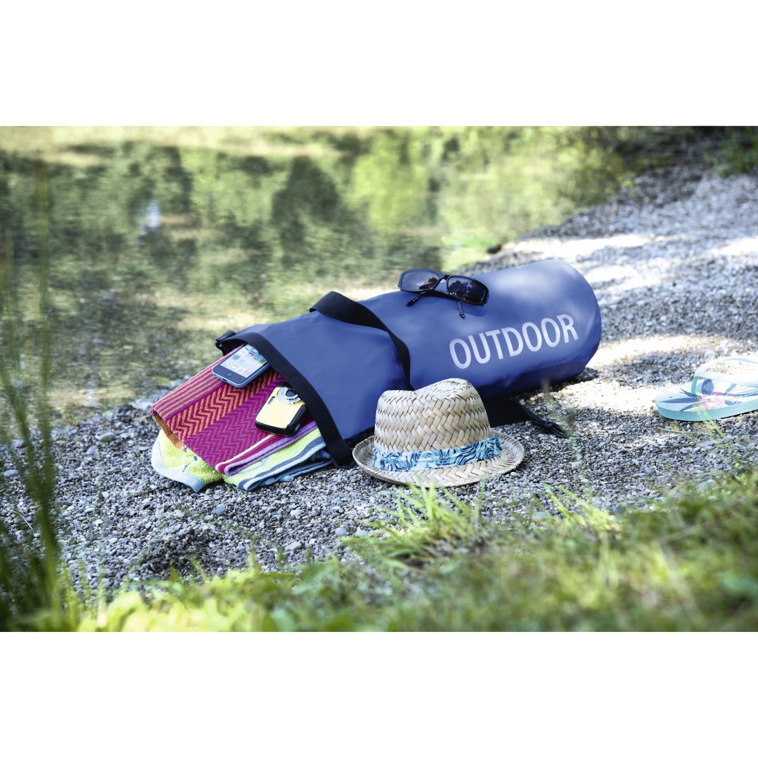 Eine blaue Tasche mit Handtüchern, Sonnenbrille, Hut und Flip-Flops am Ufer.