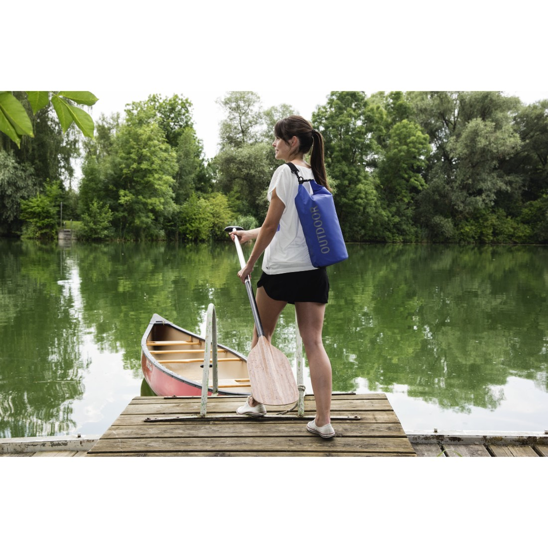 Frau mit Paddel und blauer Tasche am Steg neben Kanu.