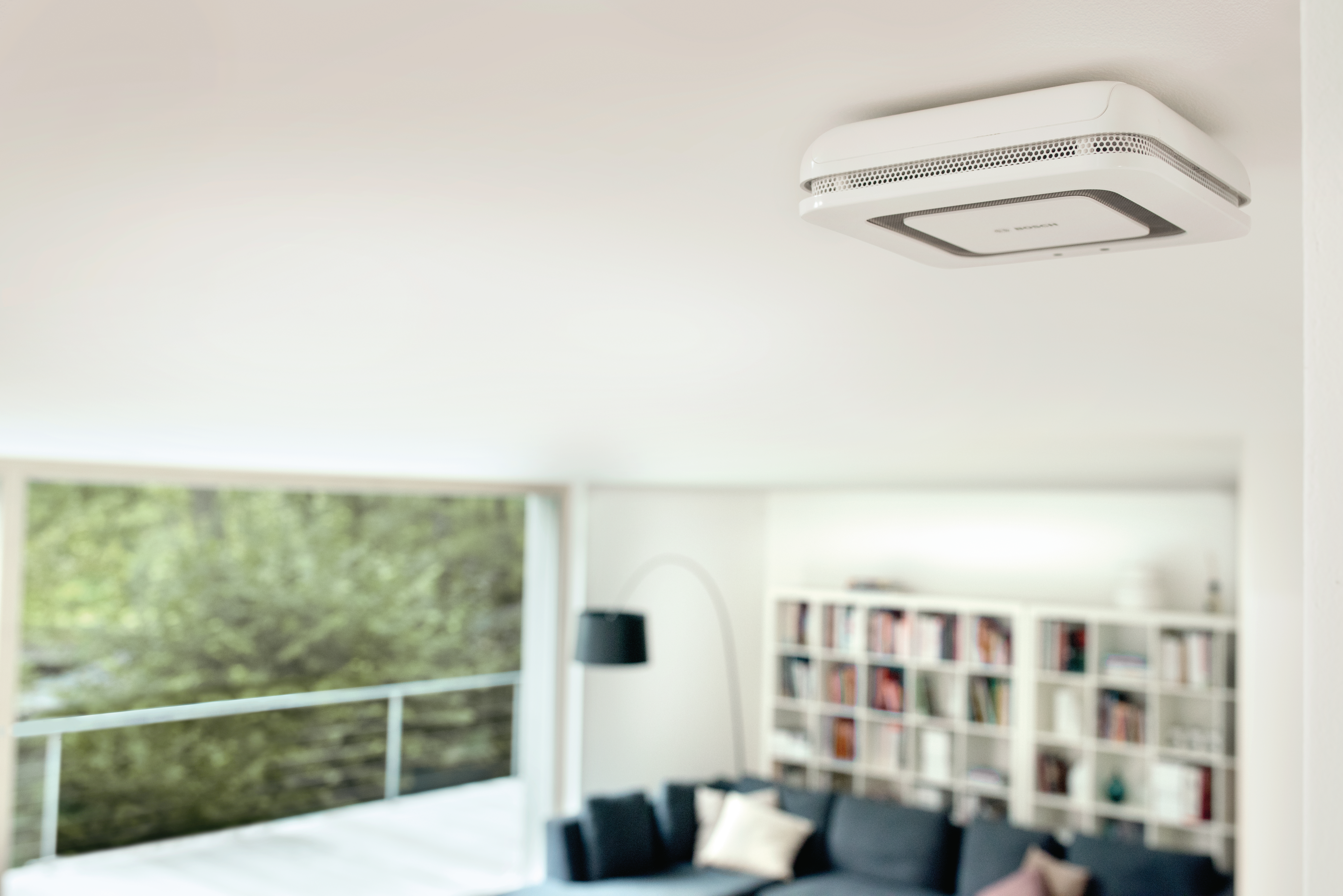 Ein weißer Rauchmelder ist an einer weißen Decke in einem Raum mit Fenster und Sofa befestigt.