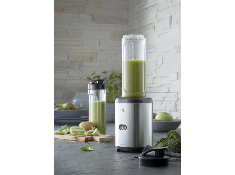 Egy ezüst smoothie mixer zöld tartalommal és gyümölcsökkel egy szürke felületen.