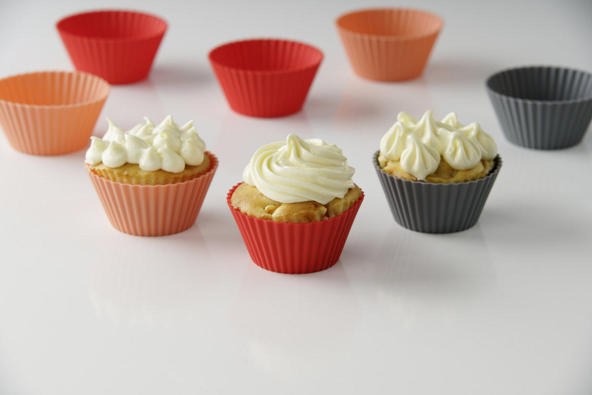 Cupcakes in roten, orangenen und grauen Silikonformen, mit weißem Frosting, auf weißer Fläche.