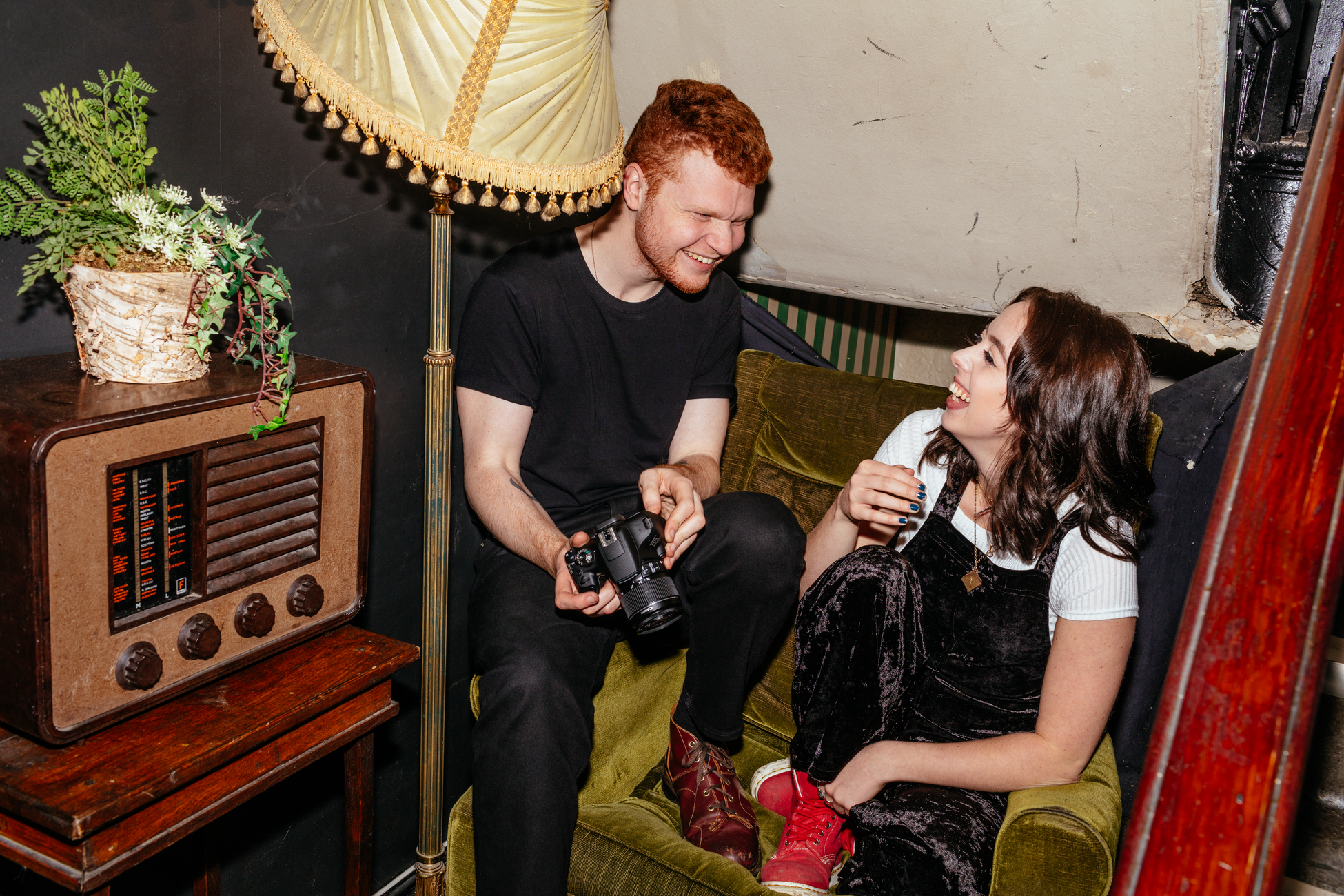 Ein Mann und eine Frau lachen auf einem grünen Sofa. Eine Kamera wird gehalten. Ein Vintage-Radio steht daneben.