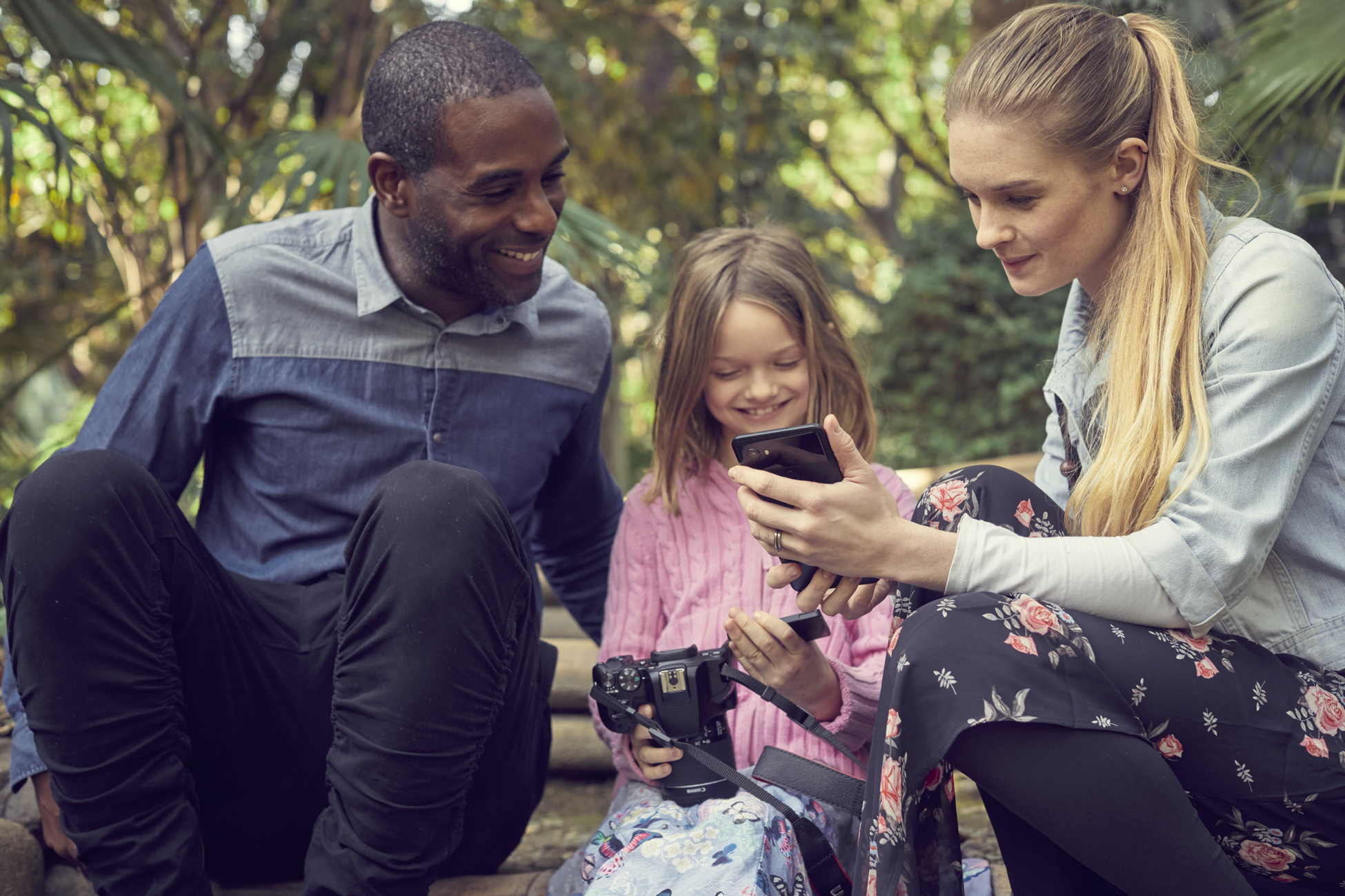 Eine dreiköpfige Familie betrachtet ein Telefon. Sie sitzen auf Stufen, umgeben von Pflanzen.