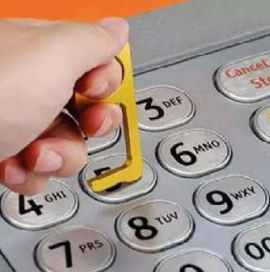 Une main utilise un outil jaune pour appuyer sur un bouton du clavier d'un guichet automatique.