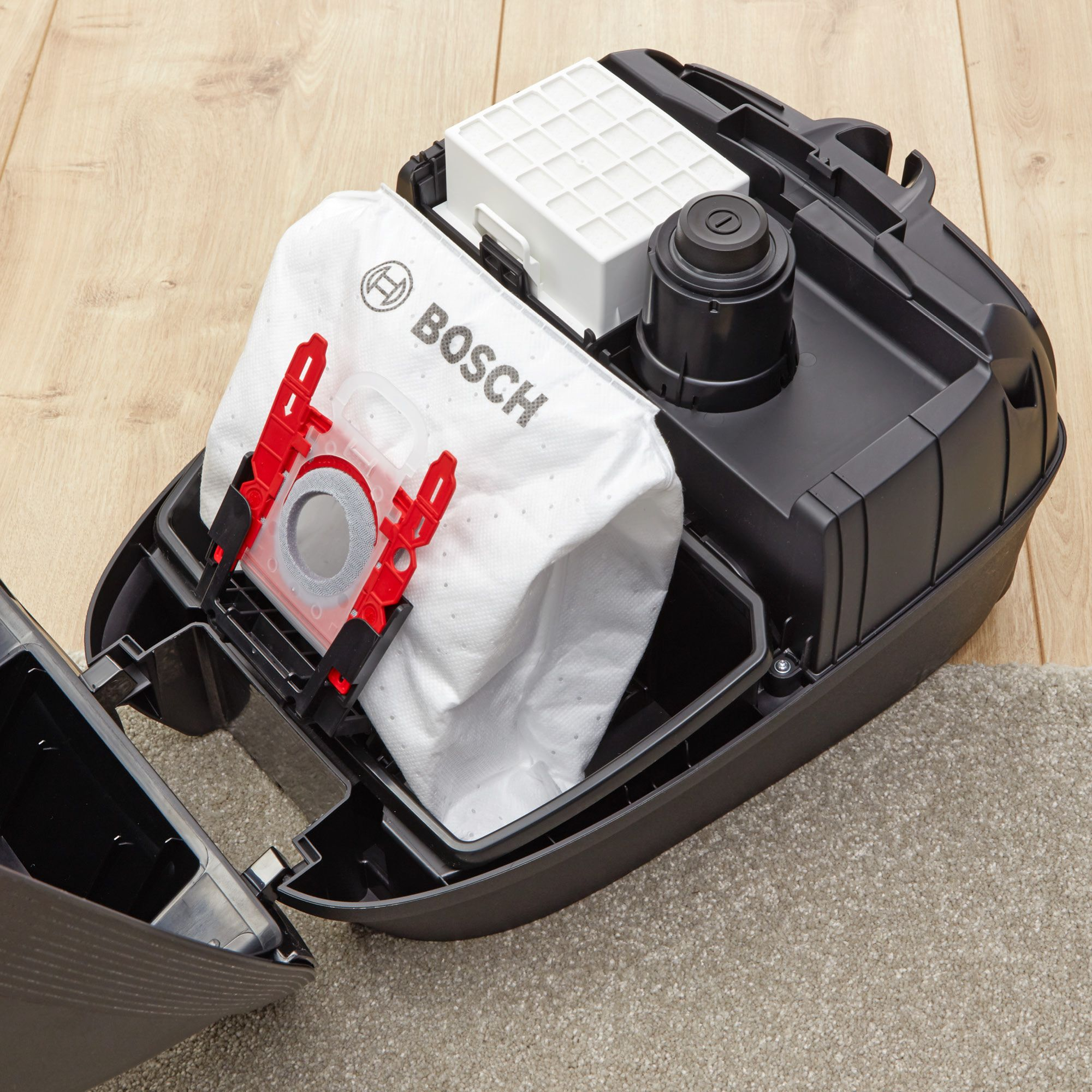 Interieur van een stofzuiger met een Bosch-zak, filter en een zwarte container op houten vloer.