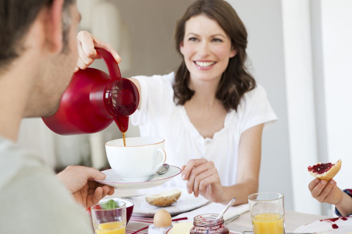 Frau gießt Kaffee in eine Tasse an einem Frühstückstisch mit zwei Personen.