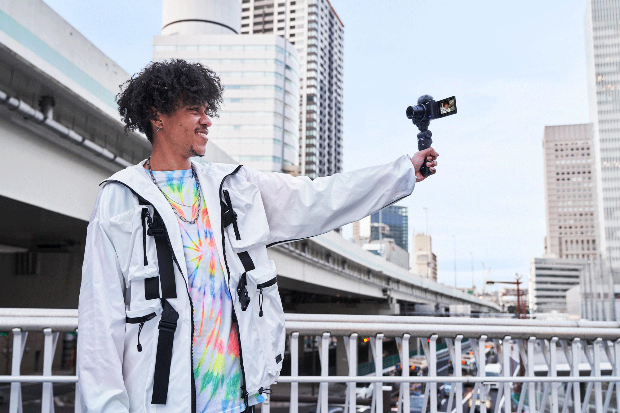 Man holding a camera, smiling. Buildings and bridge in background.