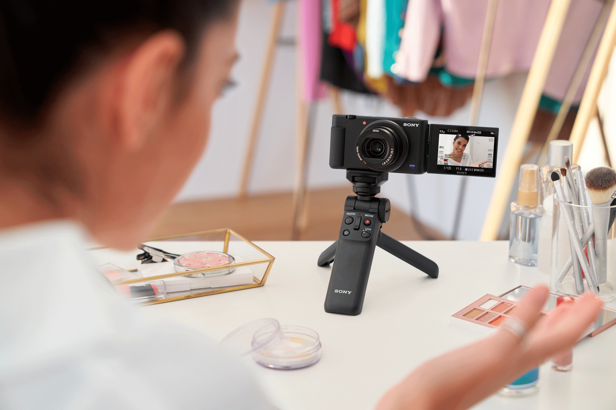 A woman is filming herself with a camera. A tripod and makeup are on the table.