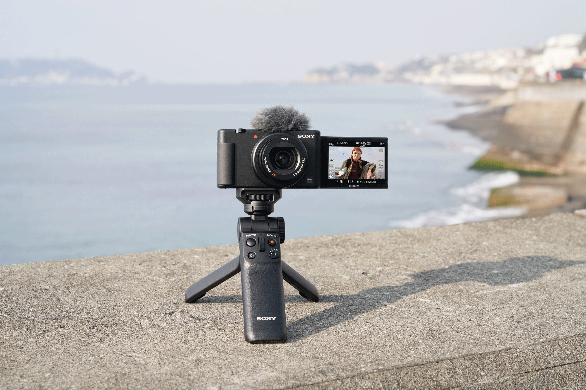 A black Sony camera on a tripod, recording a person on the screen, with a coastal background.