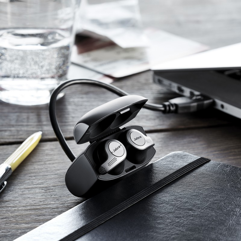 Auriculares inalámbricos en un estuche de carga sobre un escritorio de madera junto a un bolígrafo, un vaso de agua y una laptop.