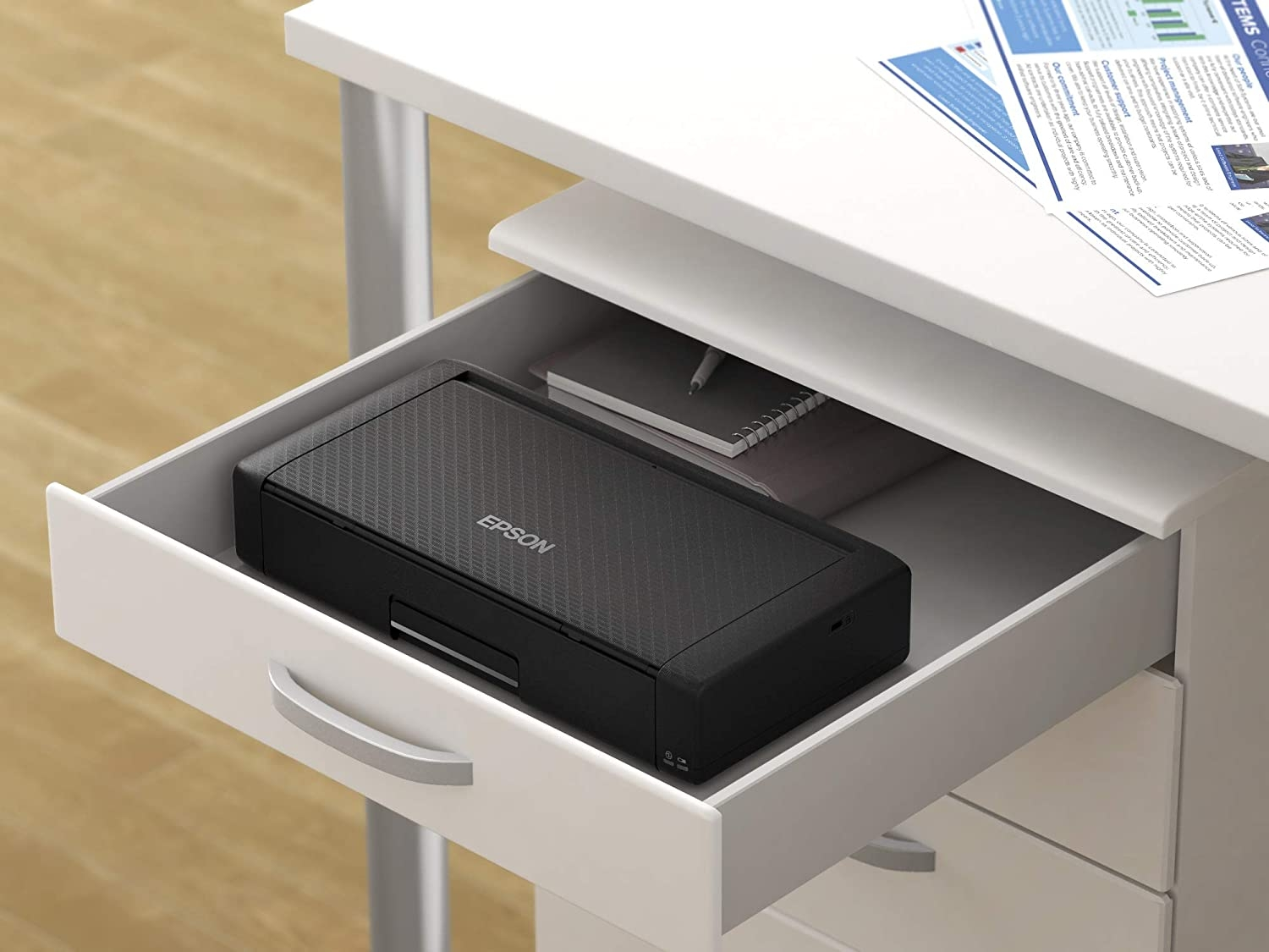 Una impresora Epson negra dentro de un cajón blanco. Un cuaderno y un bolígrafo también son visibles en el cajón.