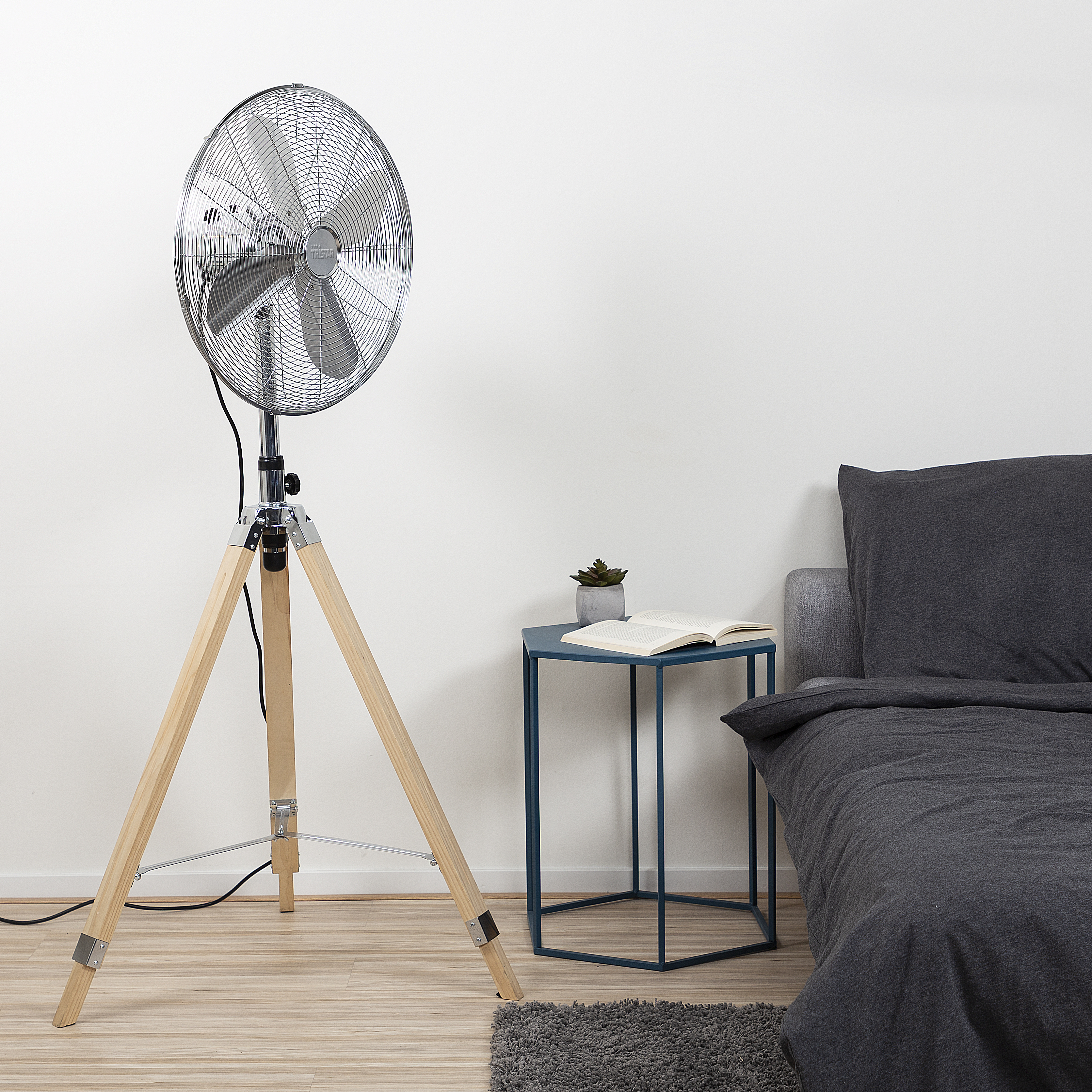 Silberner Standventilator mit Holzstativ, neben blauem Tisch und Bett.