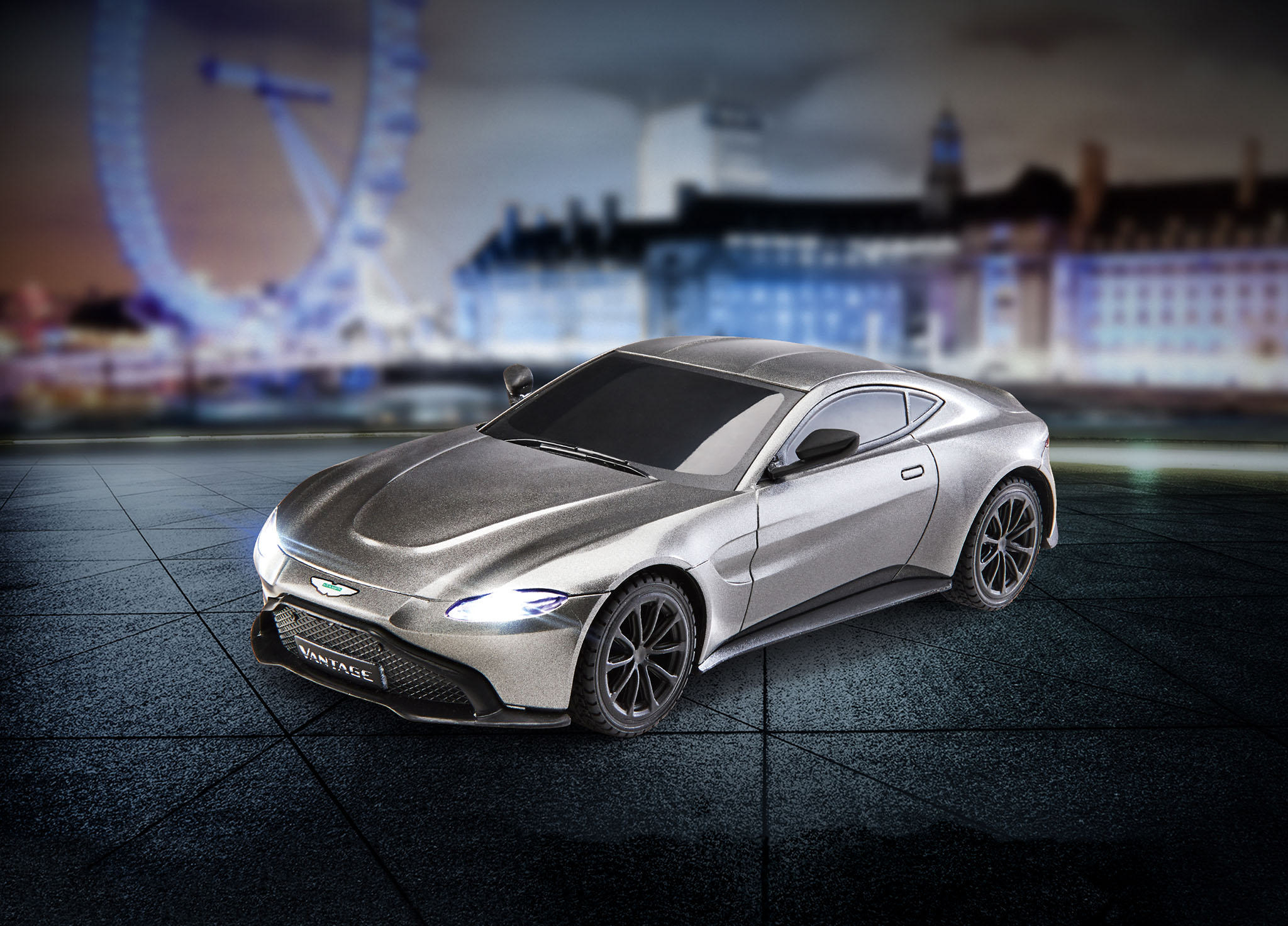 Ein graues Aston Martin Automodell auf dunkler Oberfläche mit verschwommenem Stadtbild im Hintergrund.