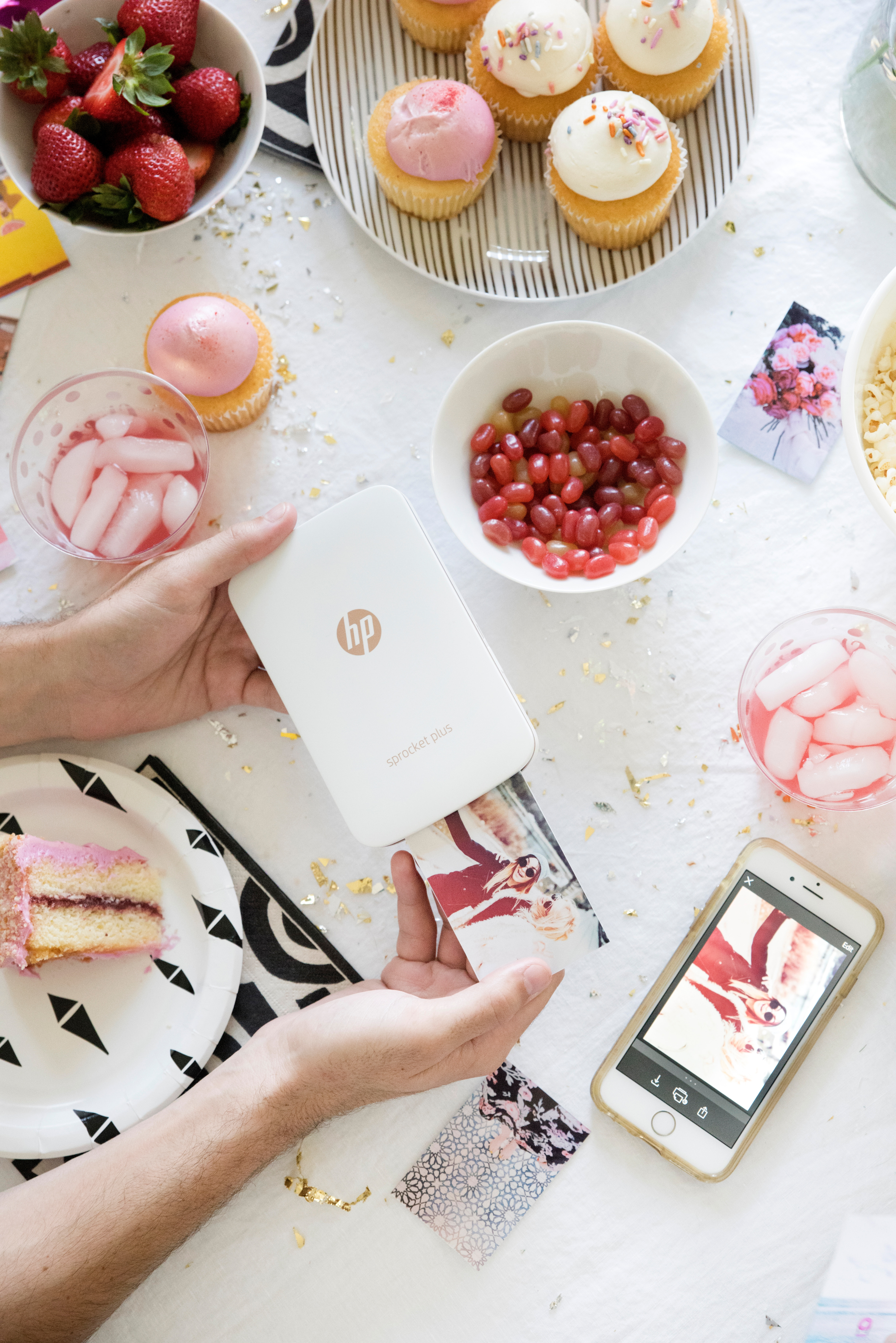 Una persona usando una impresora HP Sprocket Plus blanca rodeada de comida para fiestas y un smartphone.