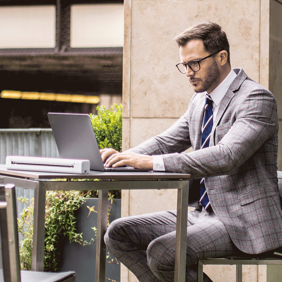 Un hombre en traje trabaja en una laptop en una mesa al aire libre con un escáner portátil.