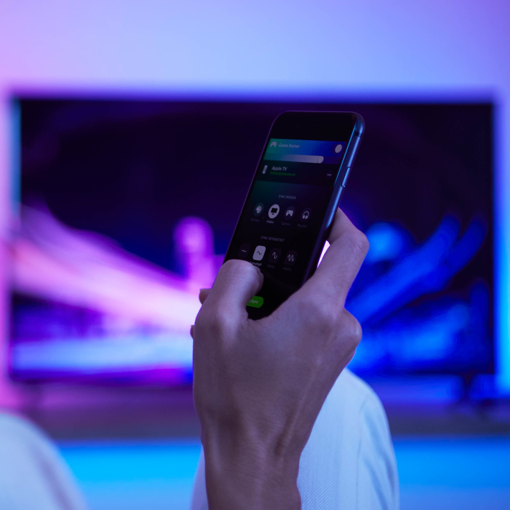 Hand die een smartphone vasthoudt met app-interface. Vage tv op de achtergrond, blauwe en paarse lichten.