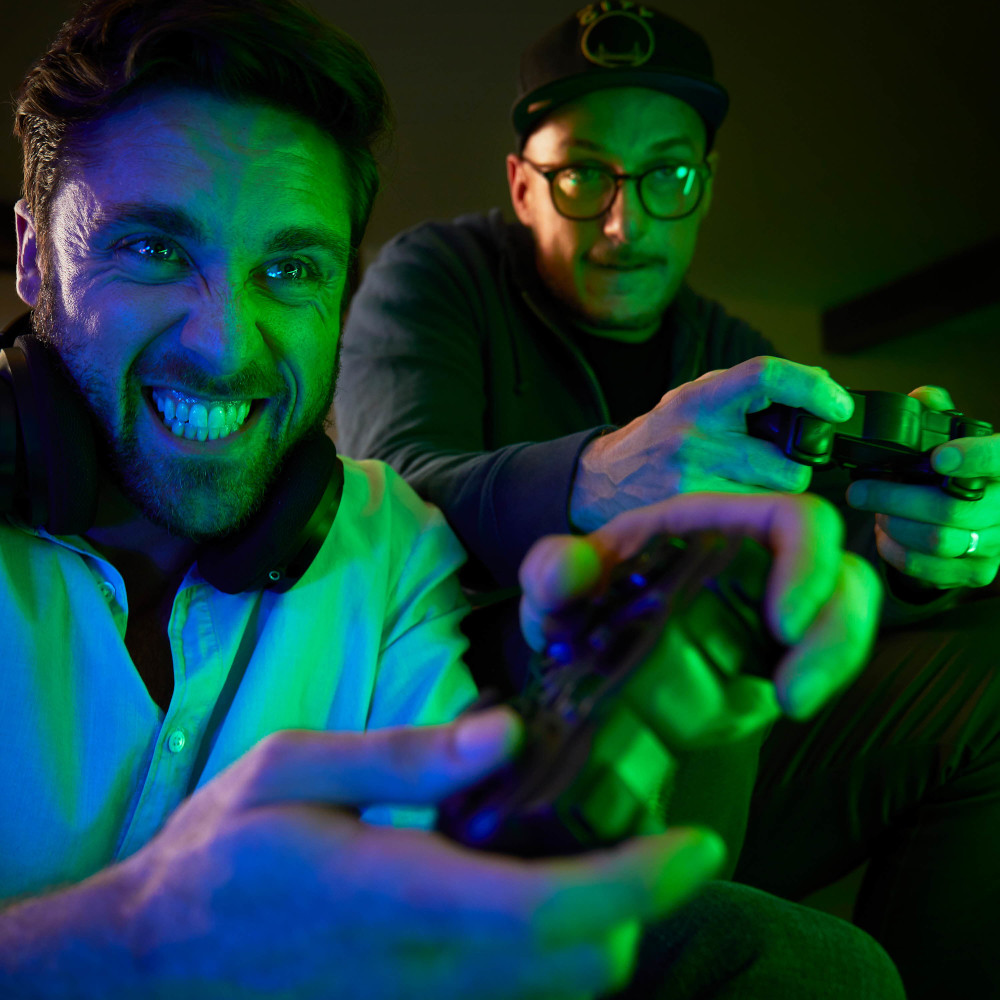 Twee mannen die videogames spelen. Eén lacht en houdt een controller vast, de ander kijkt toe.