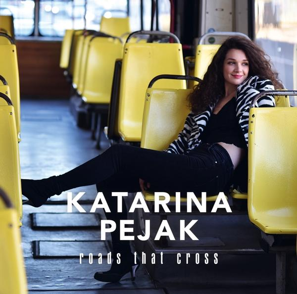 Albumcover einer Frau, die auf einem gelben Busschalter sitzt. Titel: Roads That Cross.