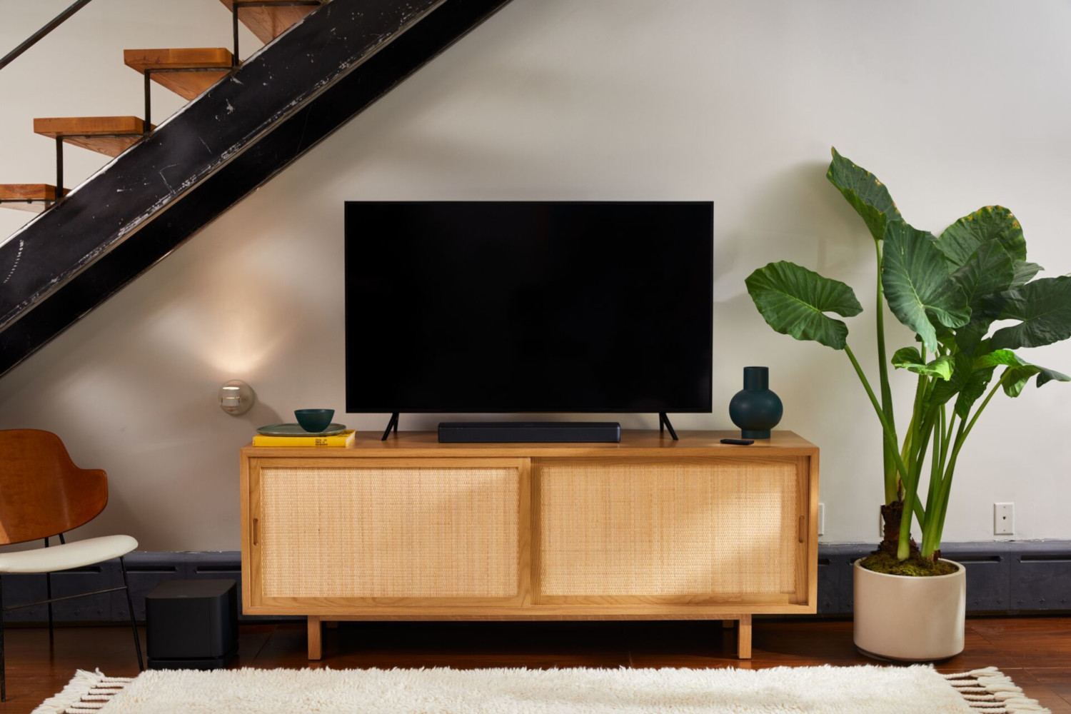 Wohnzimmer mit Fernseher auf einem Holzschrank. Treppe, Pflanze und ein weißer Teppich sind auch zu sehen.