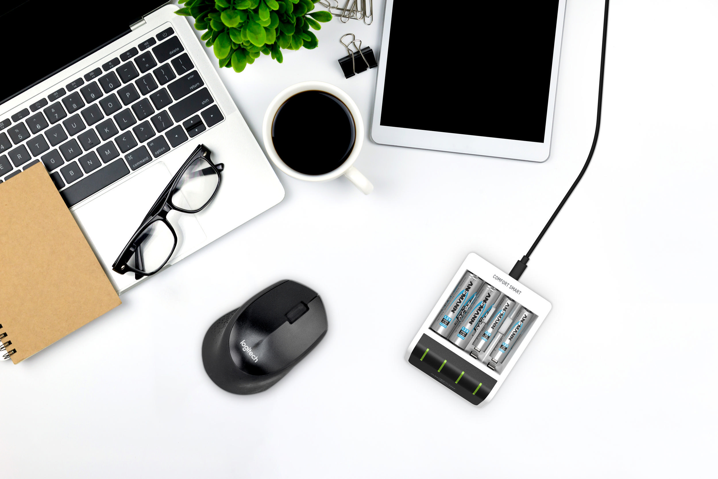 Draufsicht auf einen Schreibtisch mit Laptop, Kaffee, Brille, Maus und Ladegerät mit Batterien.