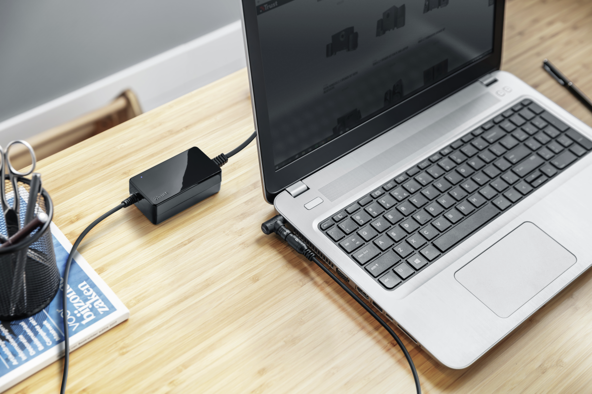 Laptop egy fekete hálózati adapterhez csatlakoztatva egy fa asztalon, egy magazin és olló mellett.
