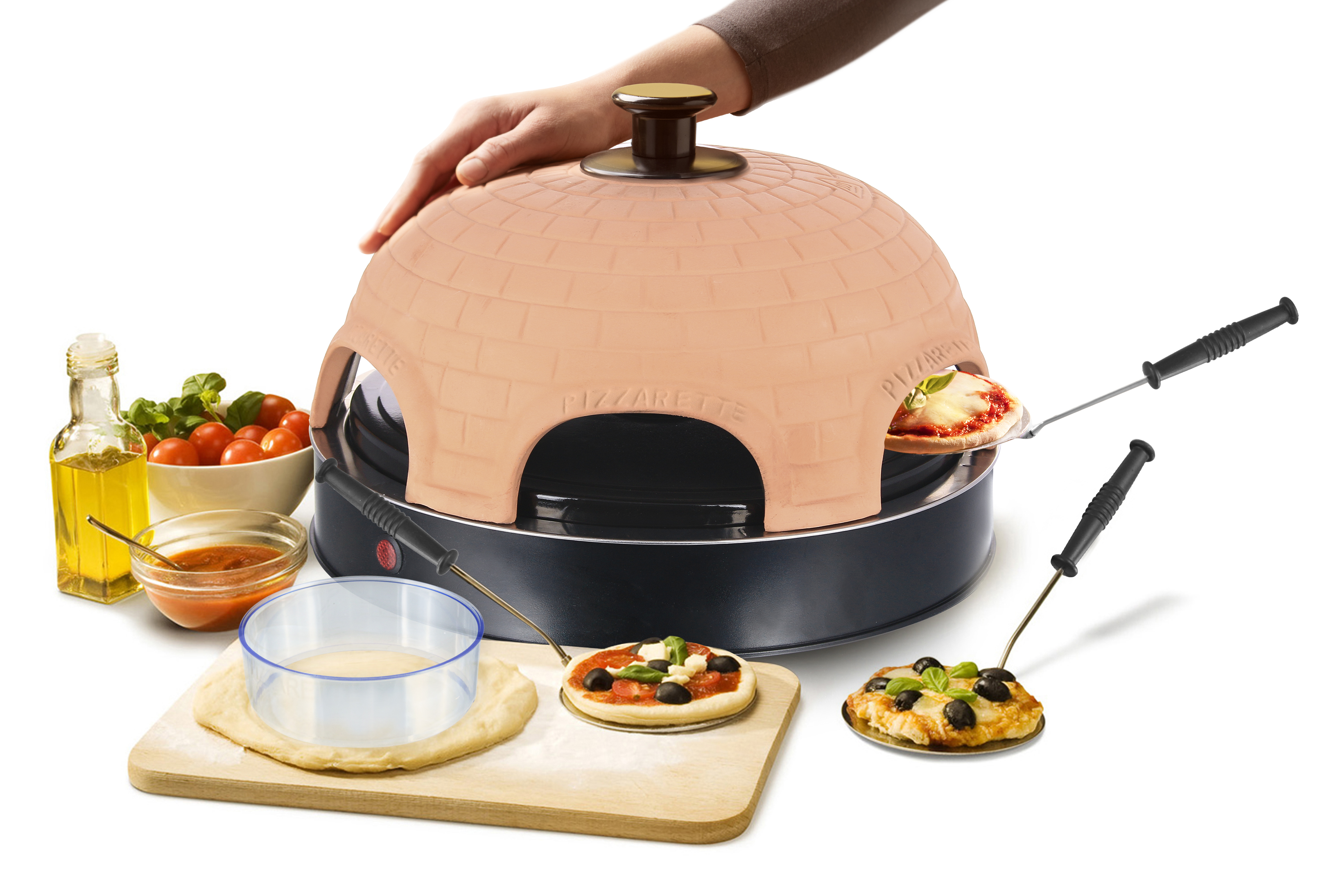 Pizzaofen mit Pizza auf Holzbrett. Zutaten neben Ofen: Öl, Tomaten, Teig und ein Messer.