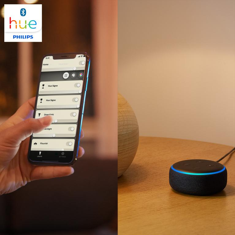 Mano de una persona operando un smartphone con controles de hogar inteligente, con un altavoz Alexa en una mesa.