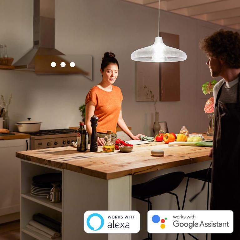 Mujer y hombre en la cocina, iluminación inteligente. Logotipos de Alexa y Google Assistant en la parte inferior.