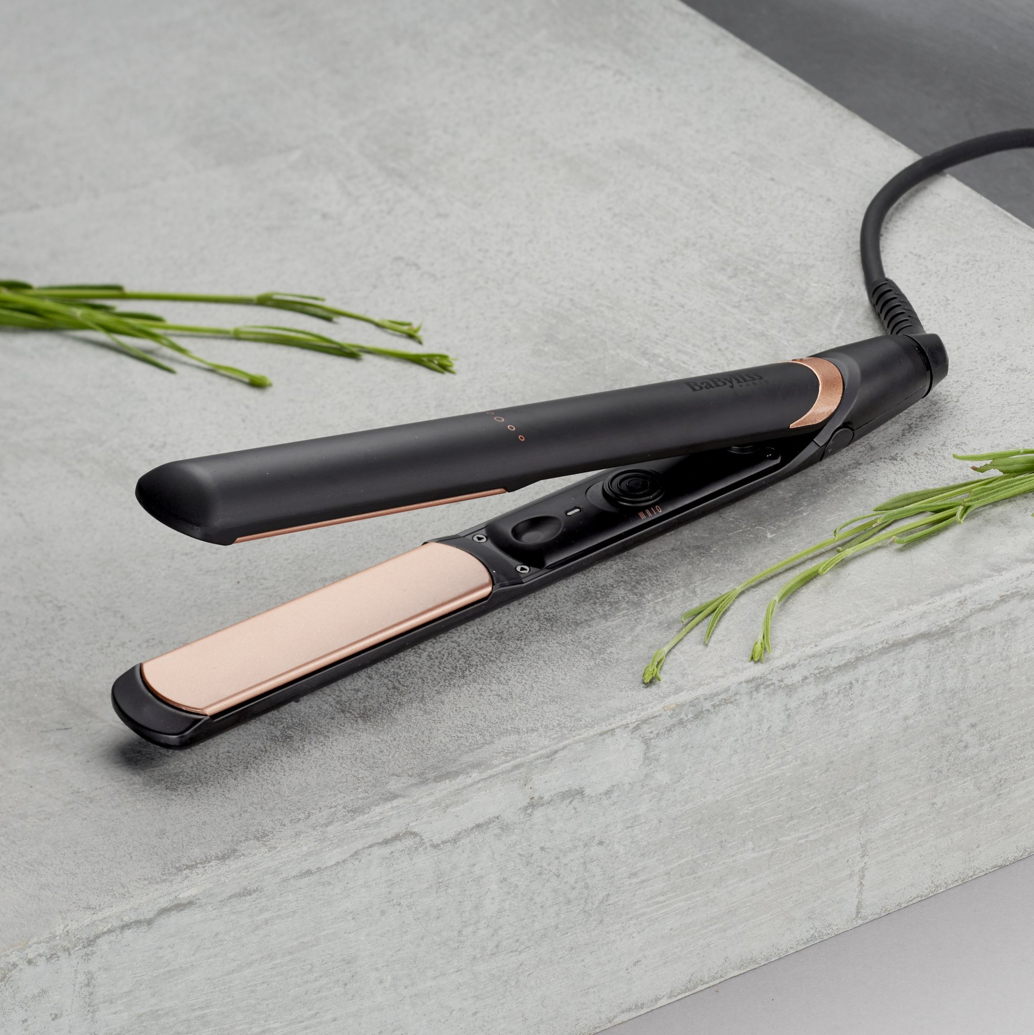 Un lisseur Babyliss noir et or rose sur une surface grise avec des brins verts.