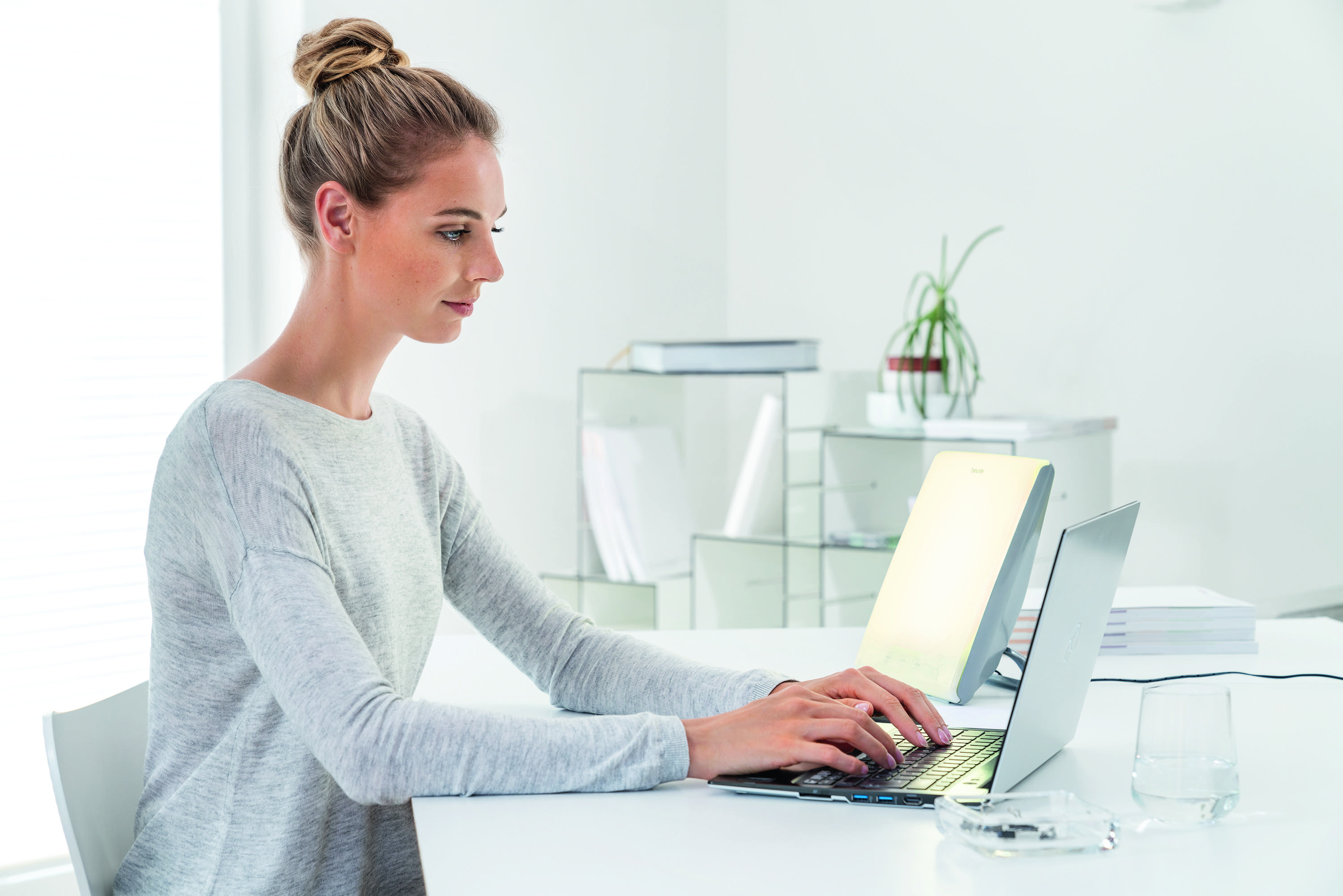 Frau arbeitet am Laptop am weißen Schreibtisch. Ein Licht und ein Glas Wasser sind vorhanden.