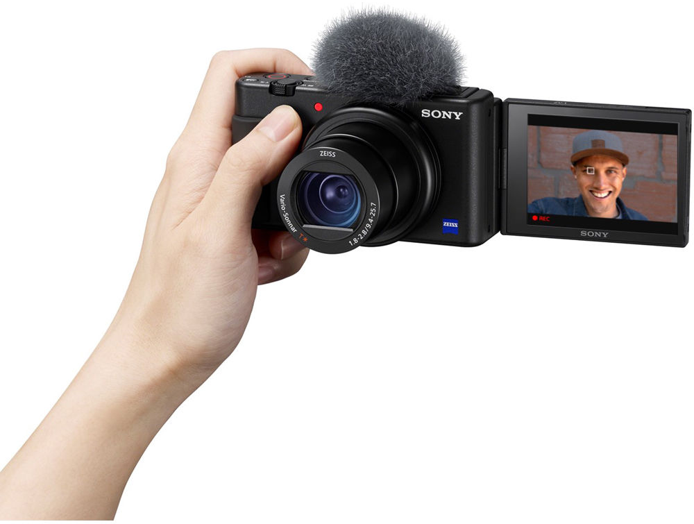 Kéz, amely egy fekete Sony kamerát tart mikrofonnal, egy személyt mutatva a képernyőn.