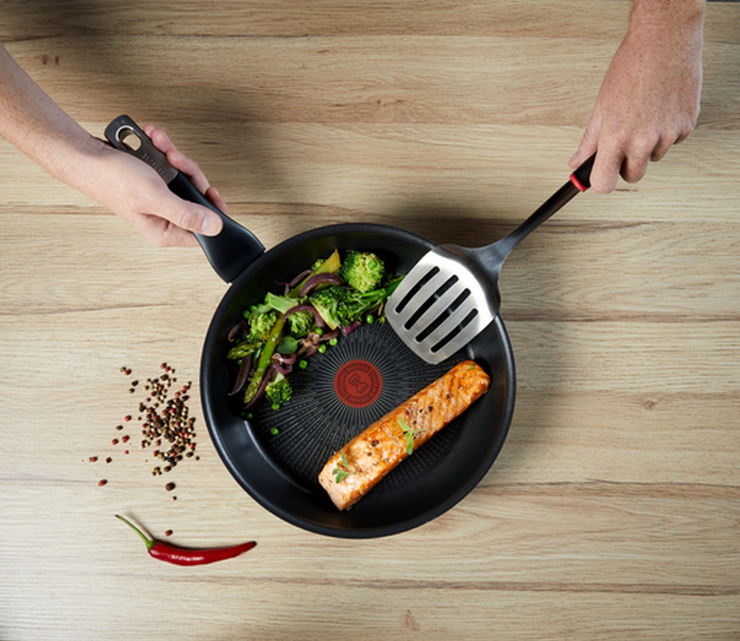 Vista cenital de una sartén con salmón cocido, verduras y una espátula sobre una mesa de madera.