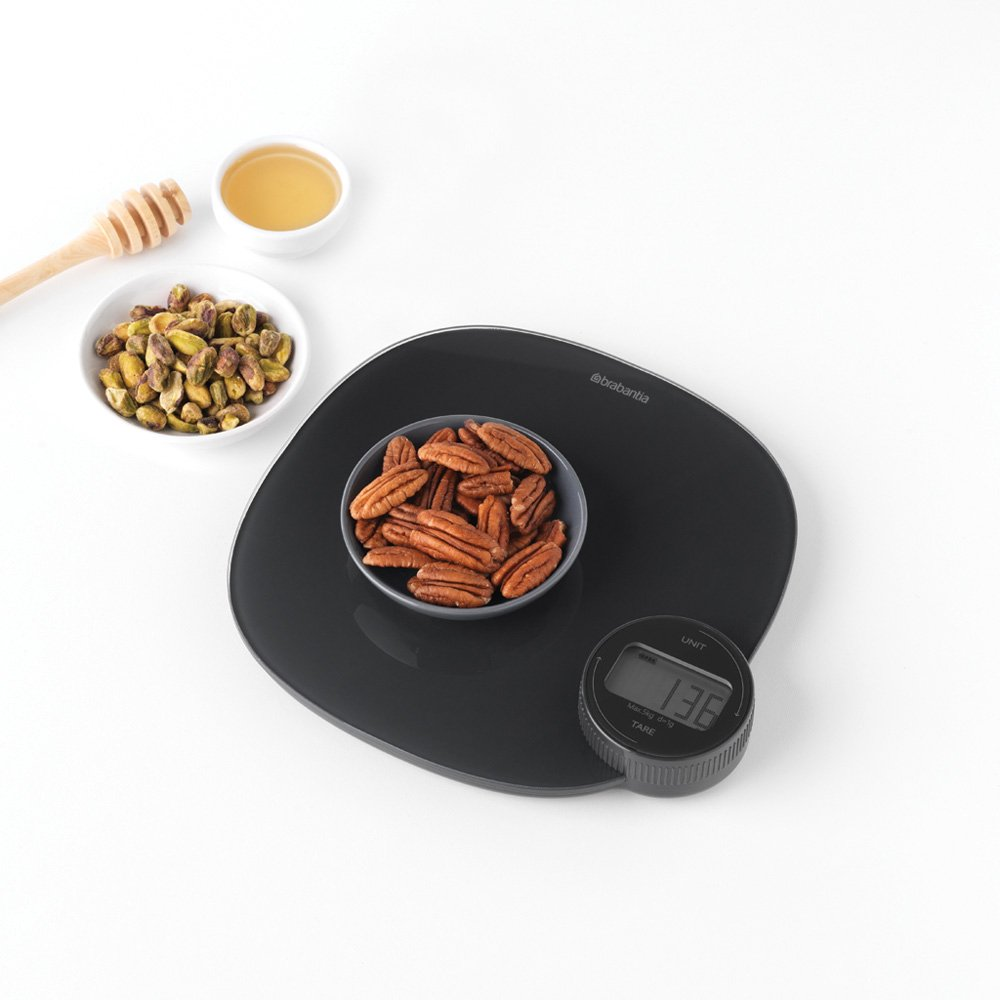 Une balance de cuisine noire avec un bol de noix de pécan, des pistaches, du miel et une baguette à miel.