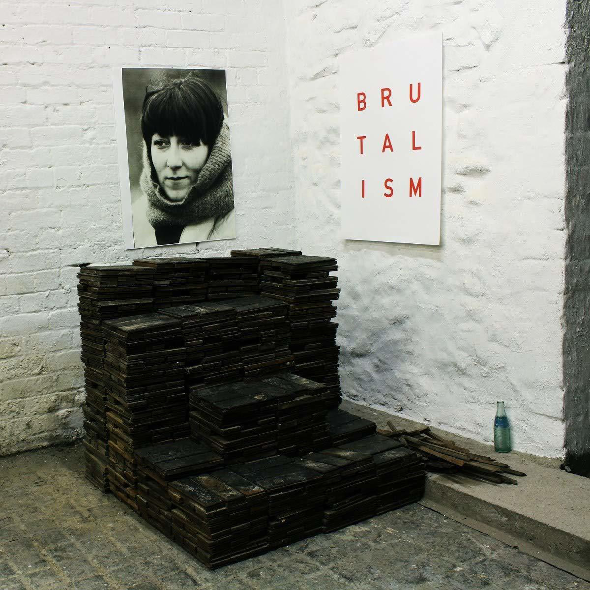 Eine Kunstinstallation mit Holzstruktur, Porträt und Schild mit der Aufschrift 'BRUTALISM'. Die Wände sind weiß.