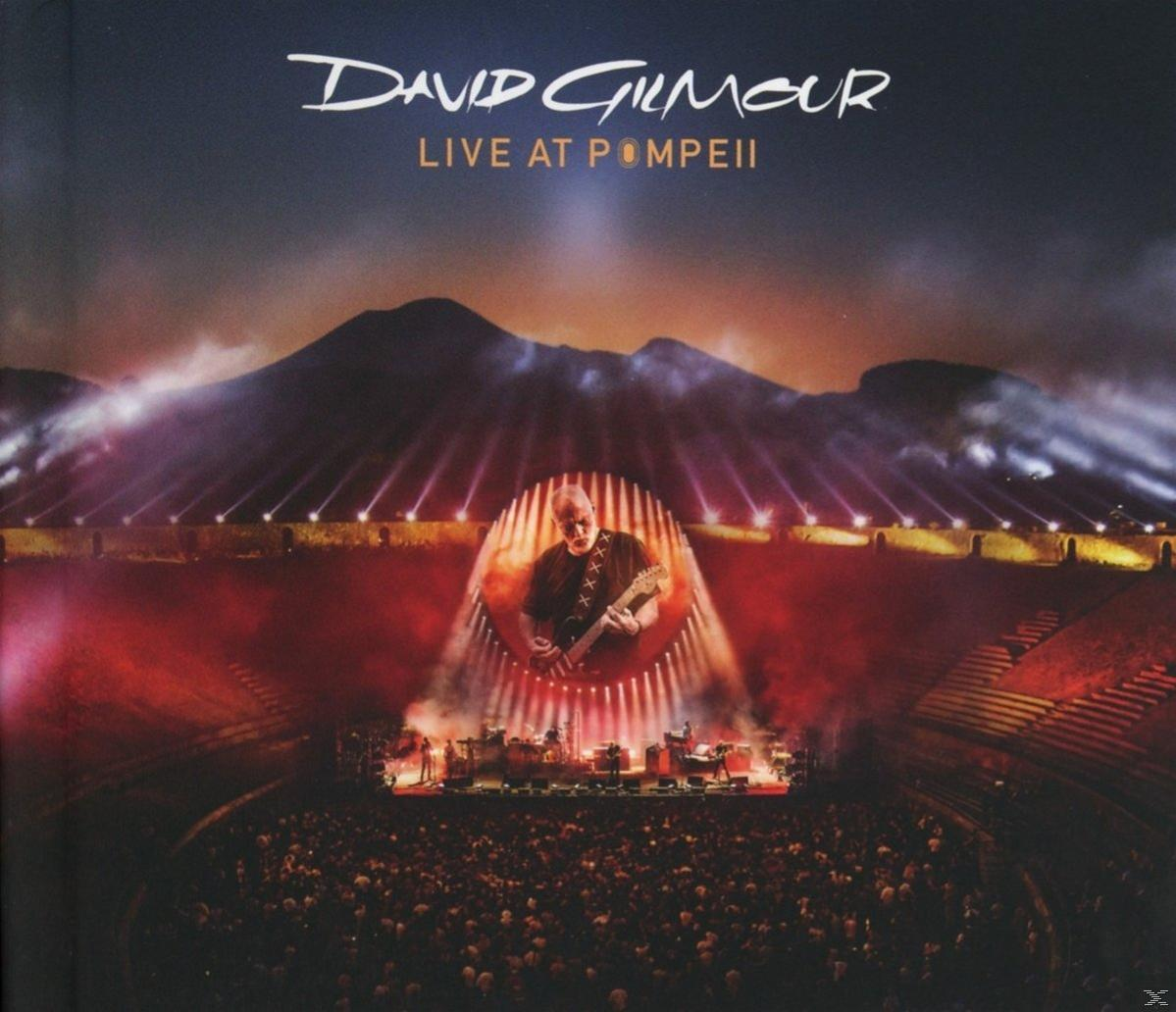 David Gilmour gitározik a színpadon a tömeg előtt. Nagy helyszín hegyi háttérrel.