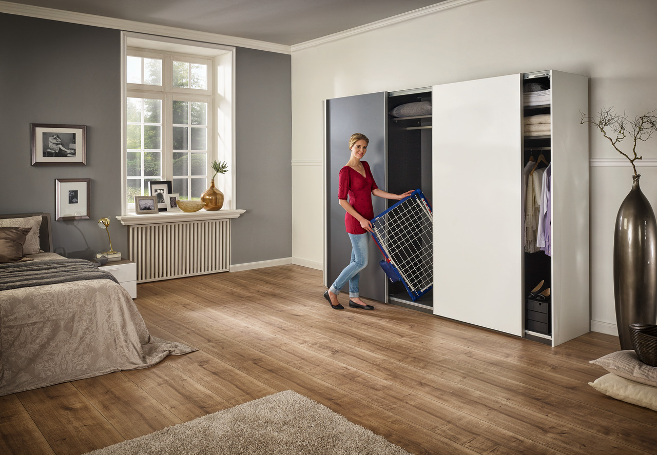 Femme en chemise rouge tient un étendoir, debout dans une chambre avec une grande armoire.