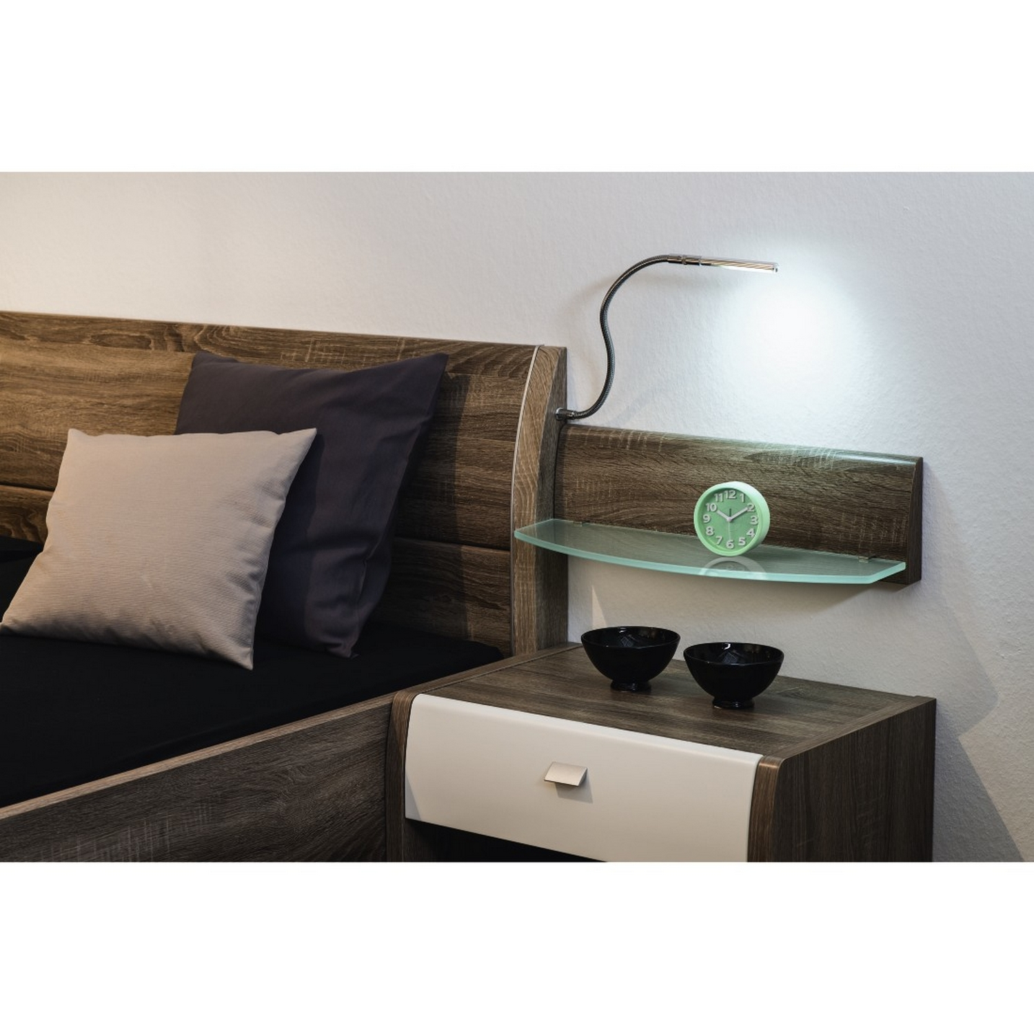 Escena de dormitorio con cama, mesita de noche, lámpara y reloj. La cama tiene almohadas.