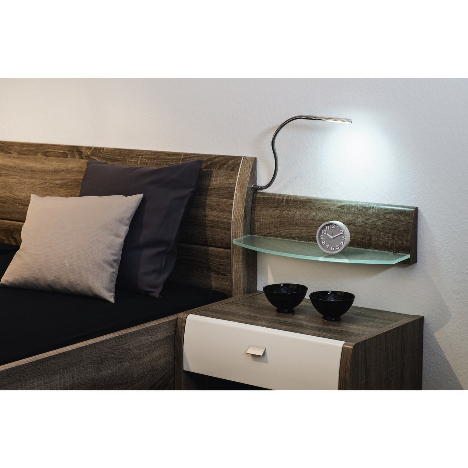 Una escena de dormitorio con una mesita de noche de madera, una lámpara, un reloj y una cama al fondo.