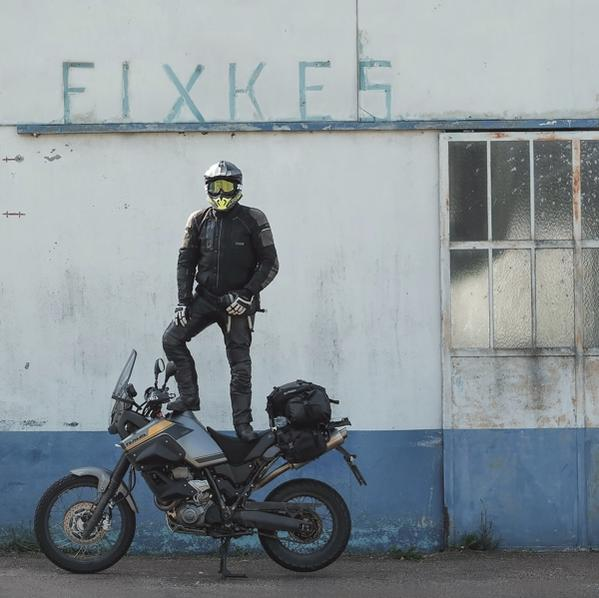 Un homme se tient sur une moto, portant un casque, devant un mur avec des graffitis.