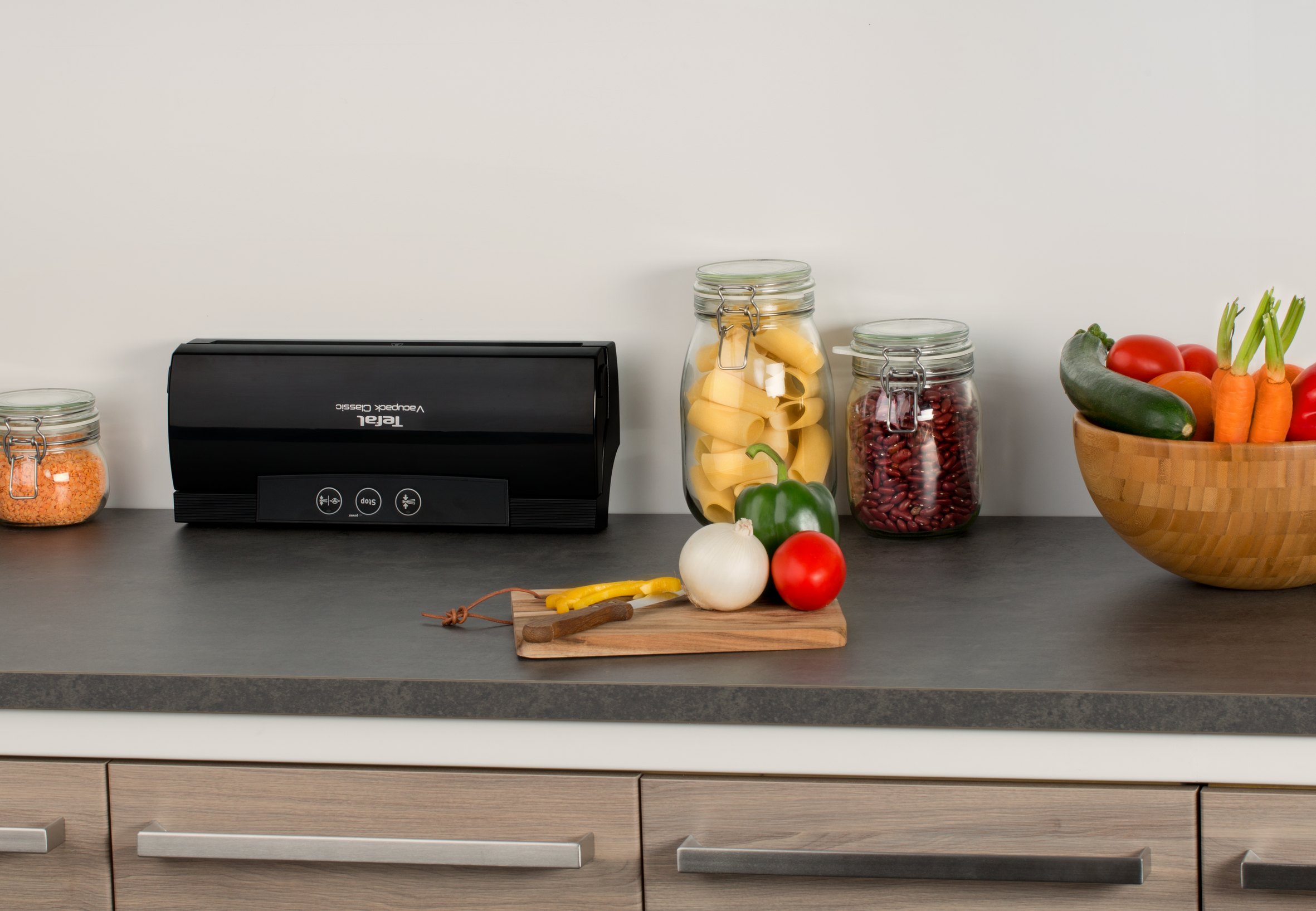 Kitchen counter with food storage containers, vegetables, cutting board and a black vacuum sealer.