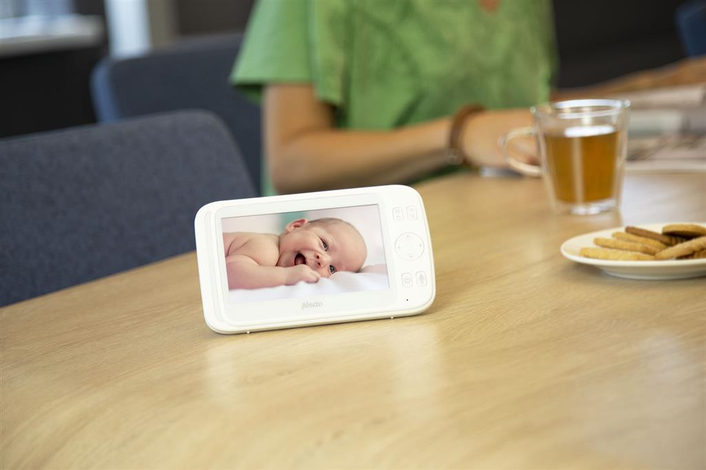 Babyphone, das ein lächelndes Baby auf einem Holztisch zeigt. Im Hintergrund ist eine Frau zu sehen.