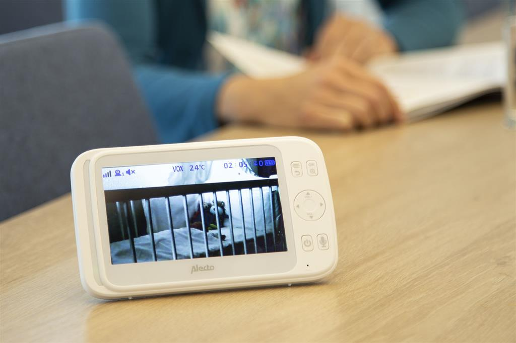 Babyphone, das ein Kinderbett mit einem Baby und Händen einer Person im Hintergrund anzeigt.