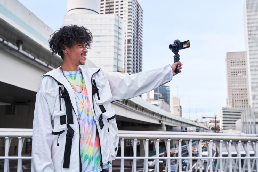 Man die een camera vasthoudt, lacht voor gebouwen. Hij draagt een witte jas en een kleurrijk shirt.