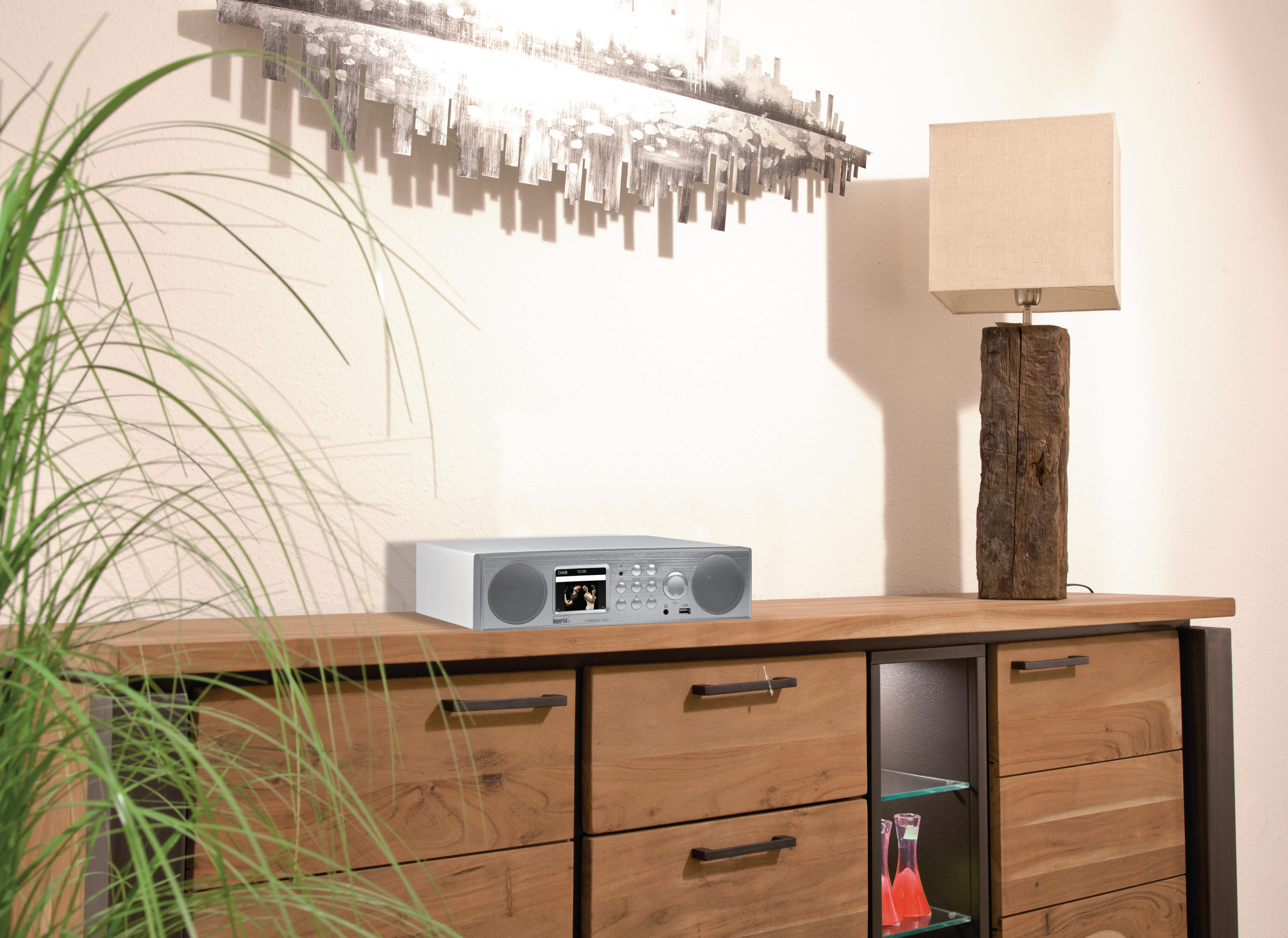 Ein silbernes Radio auf einem Holzschrank, mit Lampe und Kunst im Hintergrund, grüne Pflanze vorne.