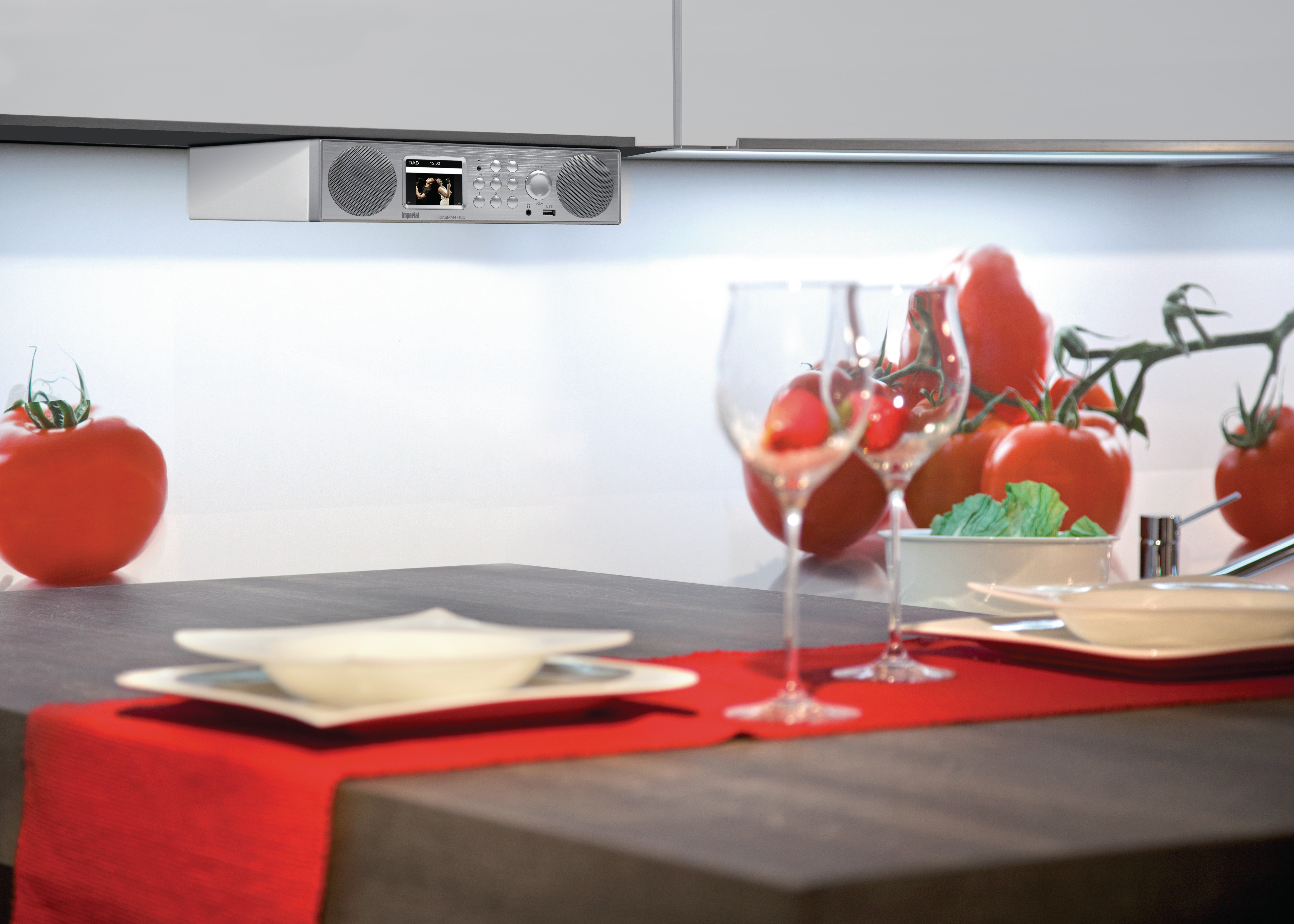 Küchenszene mit Radio, rotem Tischläufer, Tomaten und Weingläsern.