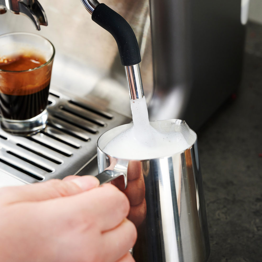 Eine Person schäumt Milch mit einer Espressomaschine in einen Metallkrug.