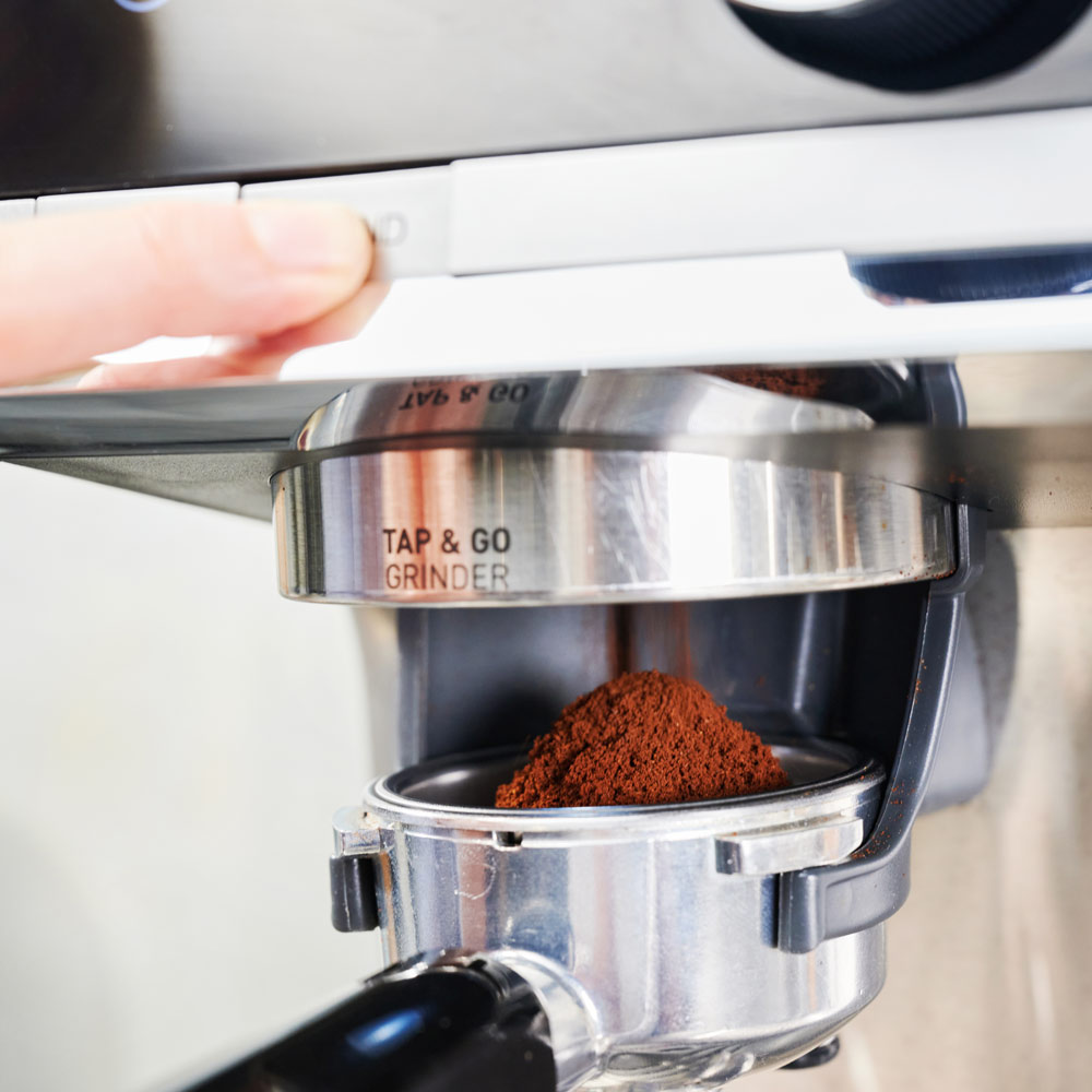 Hand drückt Knopf an Kaffeemaschine, Kaffeemehl sichtbar.