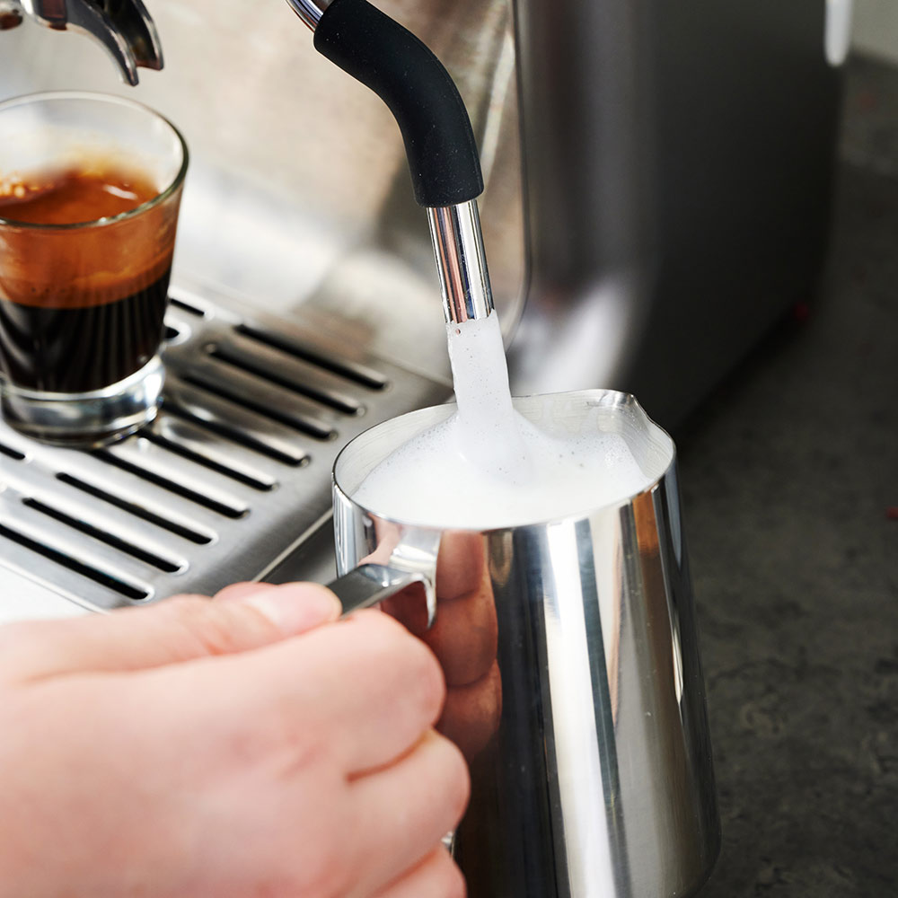 Eine Person gießt Milch in einen Metallkrug mit einer Kaffeemaschine und einem Espresso.